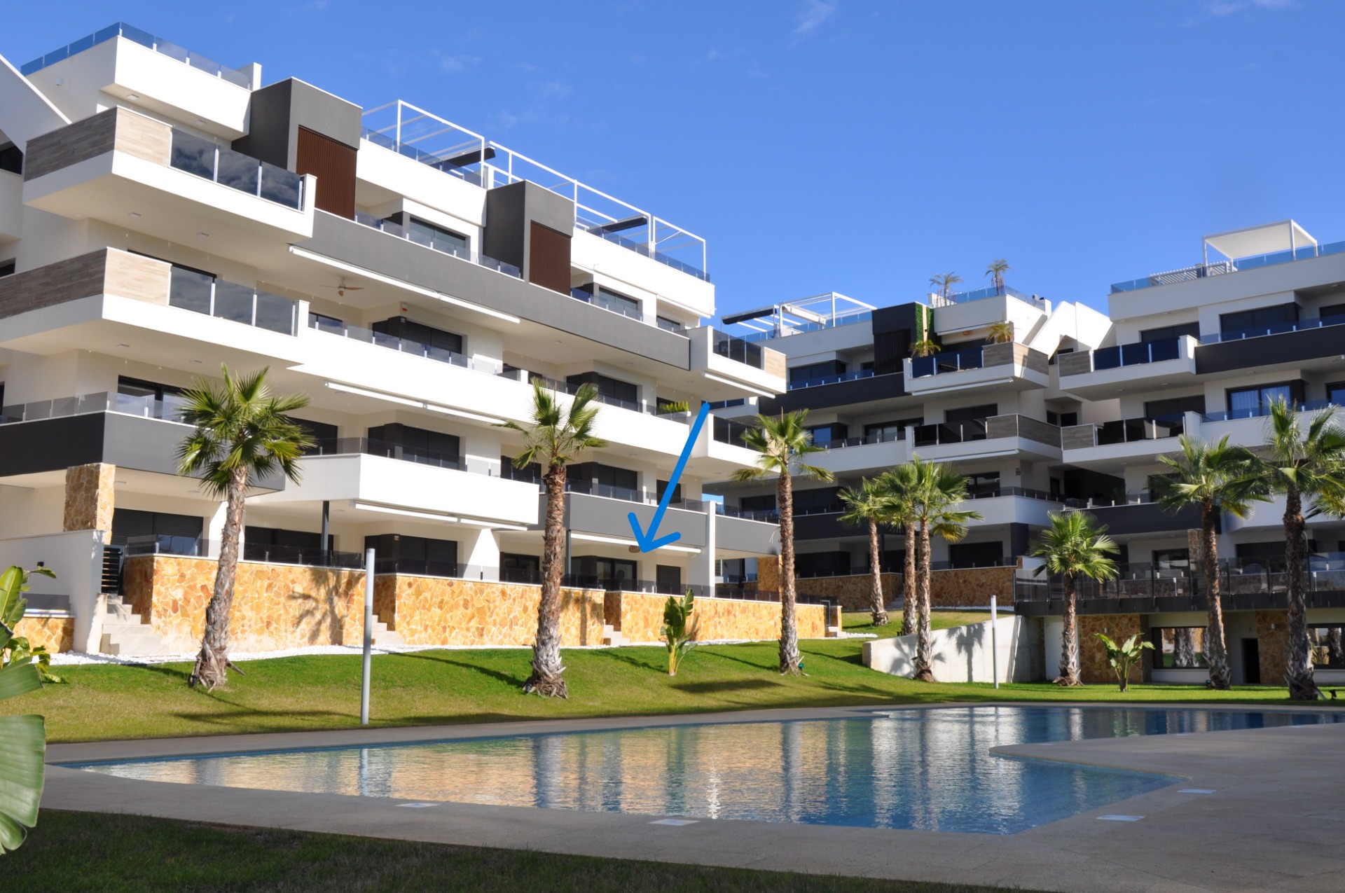 Modern appartementencomplex met balkons, palmbomen en zwembad, tegen een blauwe lucht met zonlicht.